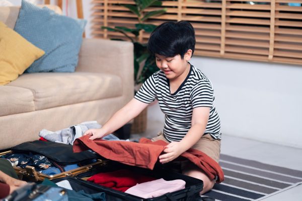 Young boy in a striped shirt kneels on a rug, packing colorful clothes into an open suitcase.