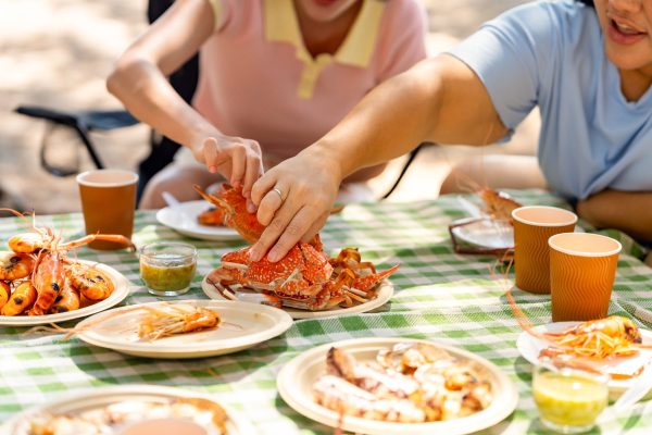 Two people crack open bright orange lobsters at an outdoor picnic on a green checkered tablecloth.