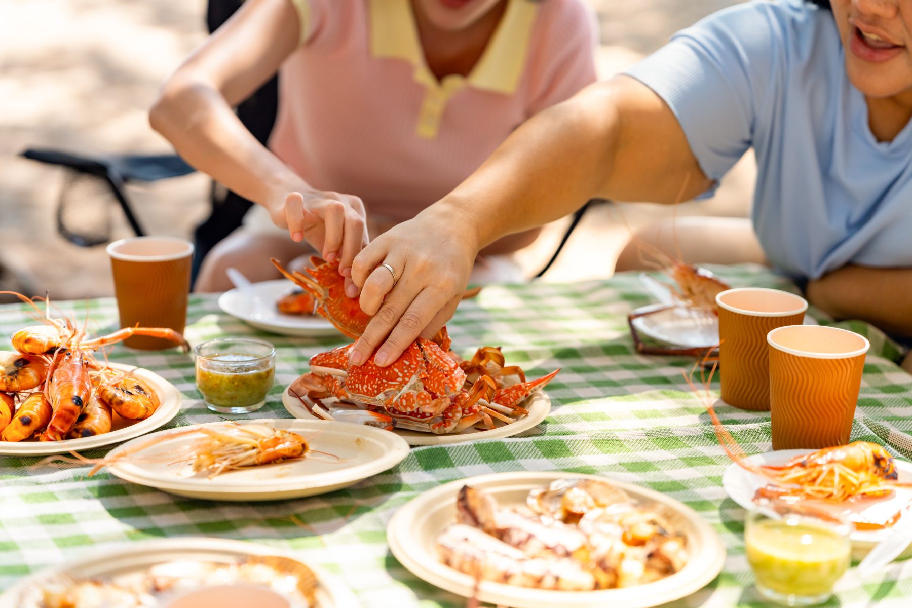 Two people crack open bright orange lobsters at an outdoor picnic on a green checkered tablecloth.