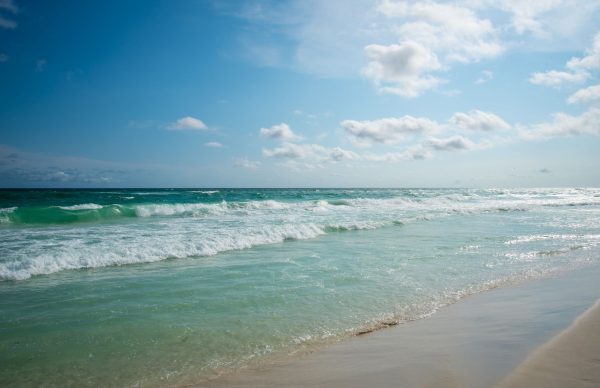 Sunlit tropical beach with turquoise waves rolling onto a pale sandy shore under a bright blue sky with few clouds.