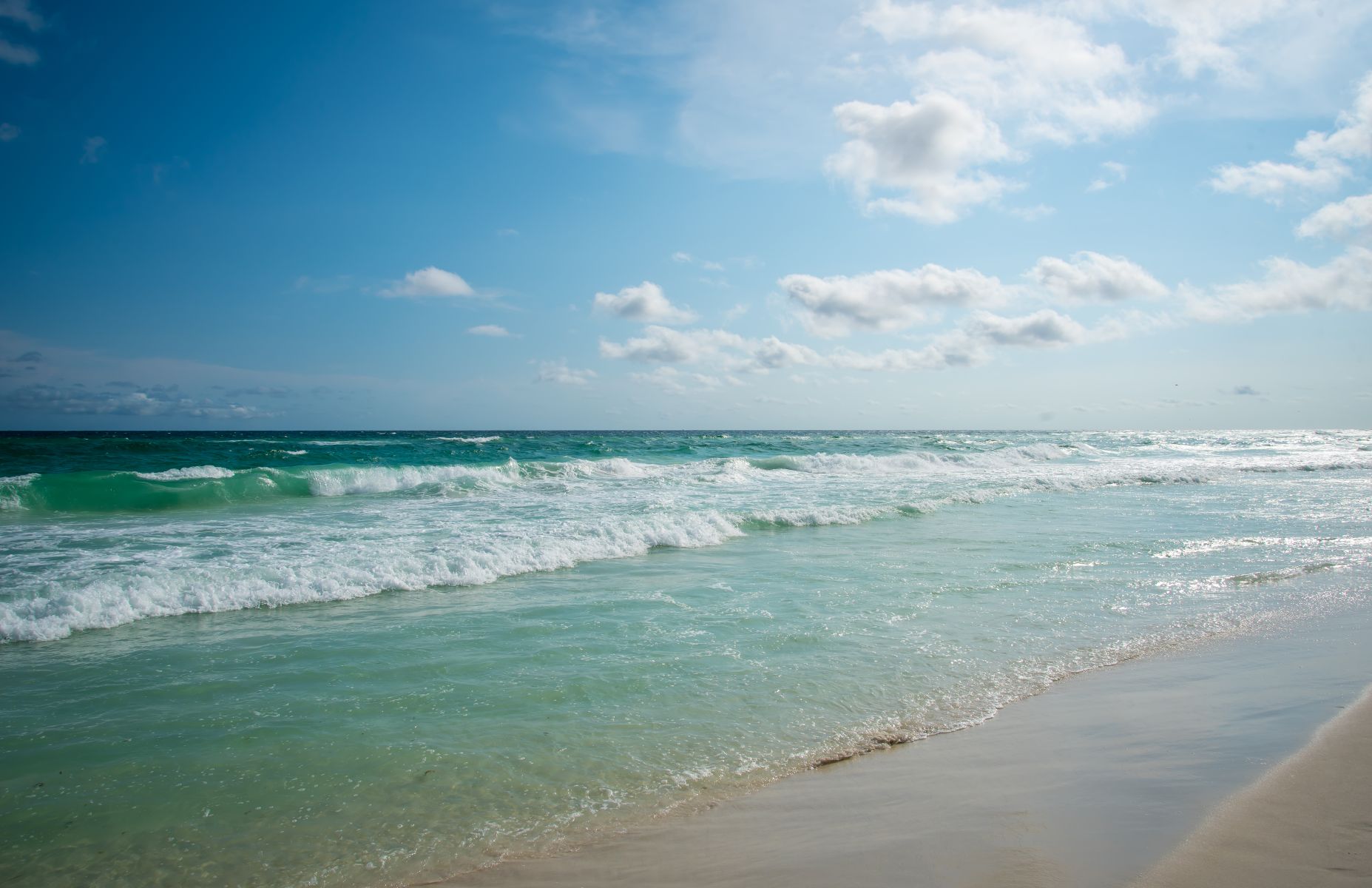 Sunlit tropical beach with turquoise waves rolling onto a pale sandy shore under a bright blue sky with few clouds.