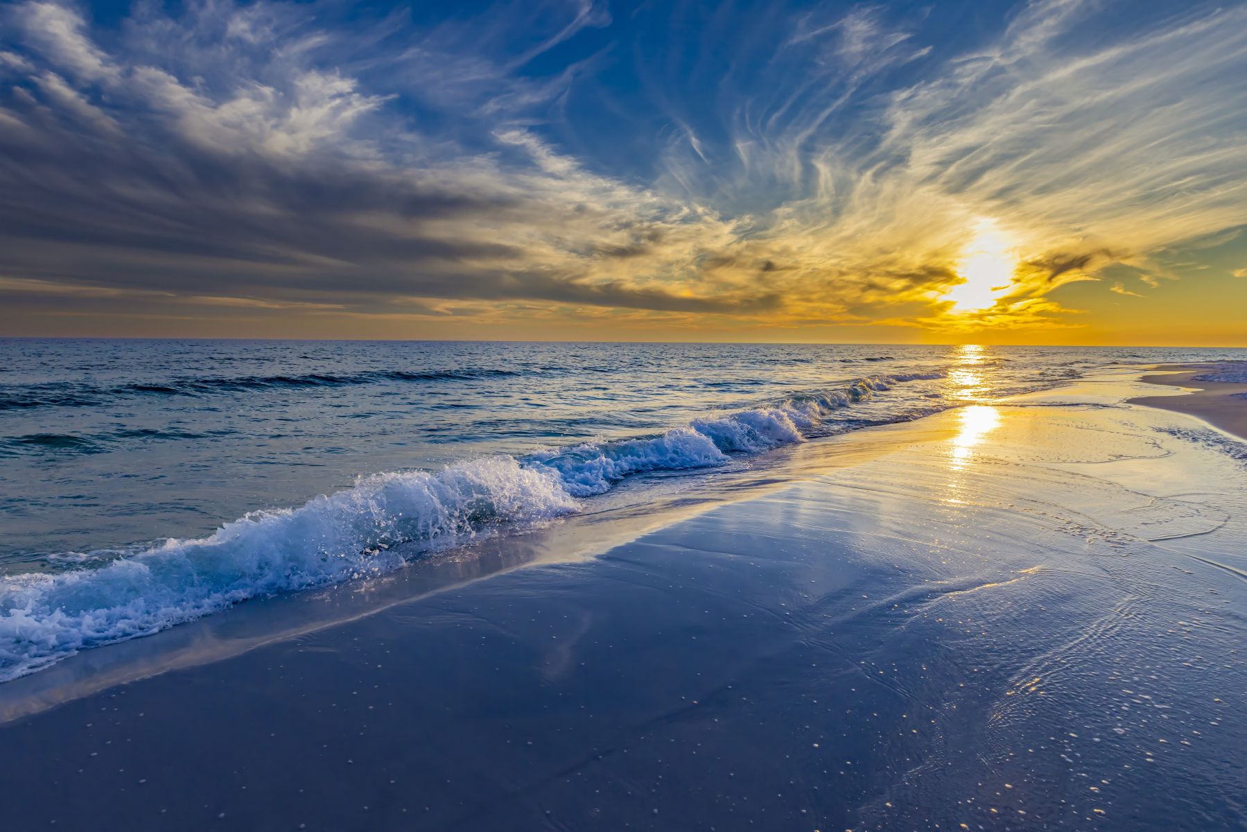 Sunset over a calm ocean with a golden sky reflecting on wet sand along a sandy beach.